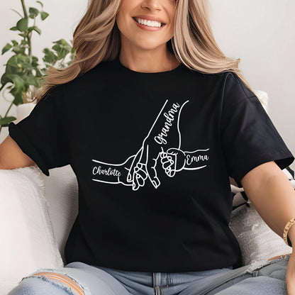 May include: Black t-shirt with a white graphic of three hands holding each other. The word "Grandma" is written in cursive on one hand, and the names "Charlotte" and "Emma" are written on the other hands.