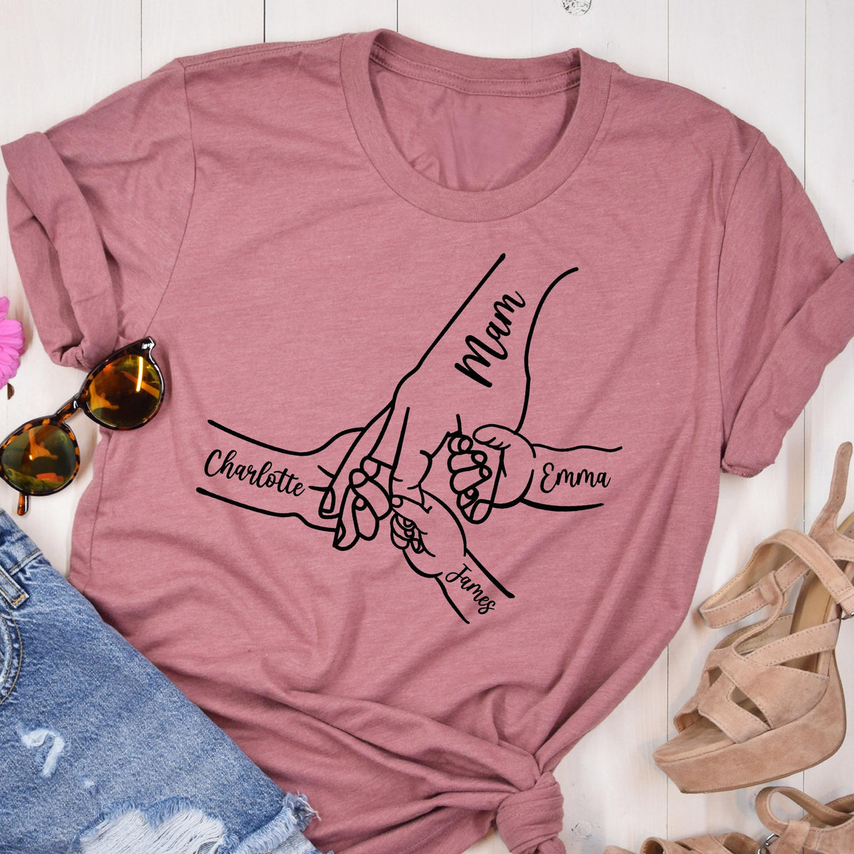May include: A dusty rose t-shirt featuring a black line drawing of hands holding each other. The word "Mam" is written on the largest hand, with the names "Charlotte", "Emma", and "James" on the smaller hands. The shirt is folded.
