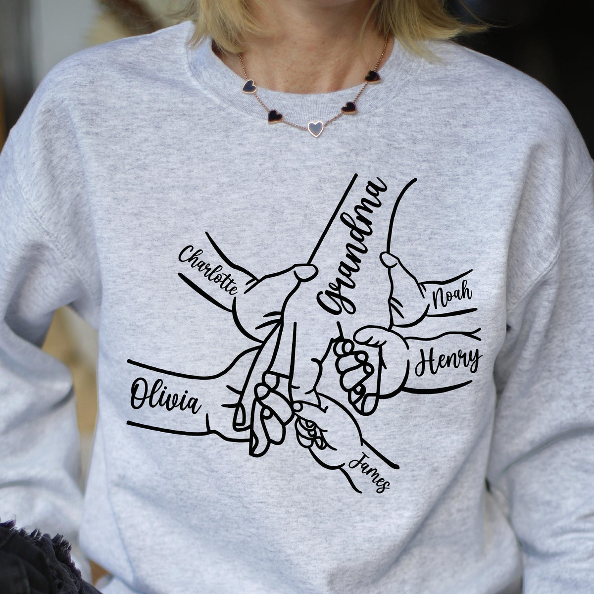 May include: A light gray sweatshirt featuring a black line drawing of hands clasped together. The word "Grandma" is written in cursive, with the names "Charlotte", "Olivia", "Noah", "Henry", and "James" also written in cursive.
