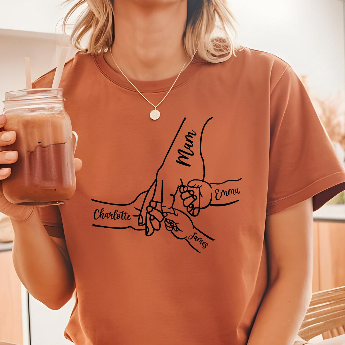 May include: A rust-colored short-sleeve t-shirt featuring a black line art graphic of multiple hands holding fingers together. The text Mam, Charlotte, Emma, and James is printed in a cursive script alongside the corresponding hand illustrations.
