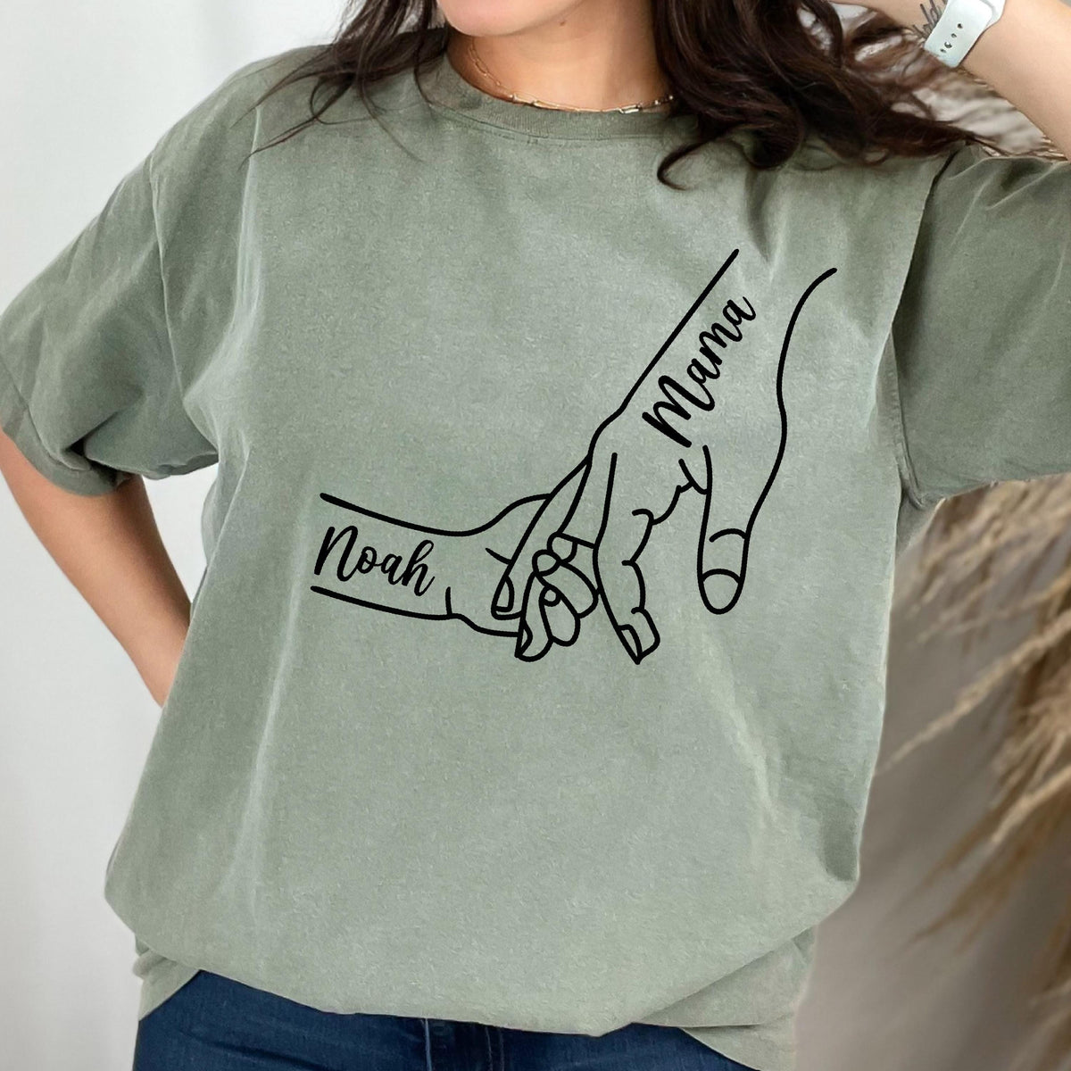 May include: A sage green short-sleeve t-shirt featuring a black line art graphic of two hands holding each other. The larger hand has the word Mama written along the fingers, and the smaller hand has the name Noah written across the wrist.