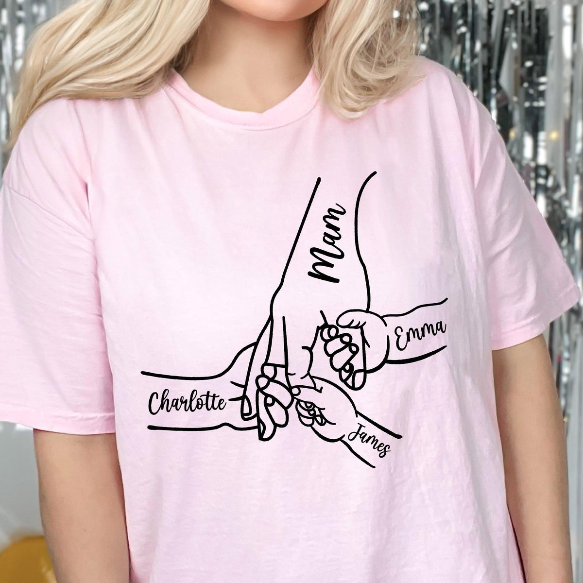 May include: A light pink t-shirt featuring a black line art graphic of one large hand holding two smaller hands. The text Mam is written on the large hand, while the names Charlotte, Emma, and James are printed near the smaller hands.