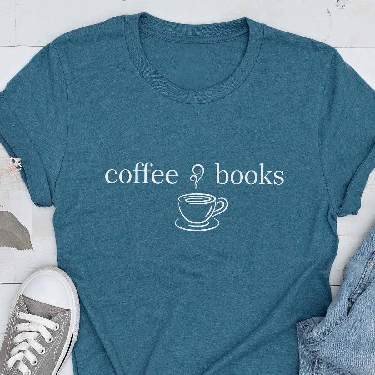 May include: Teal t-shirt with the words "coffee & books" and a coffee cup graphic in white. The shirt is made of a soft material and has short sleeves. A gray sneaker and denim are partially visible.