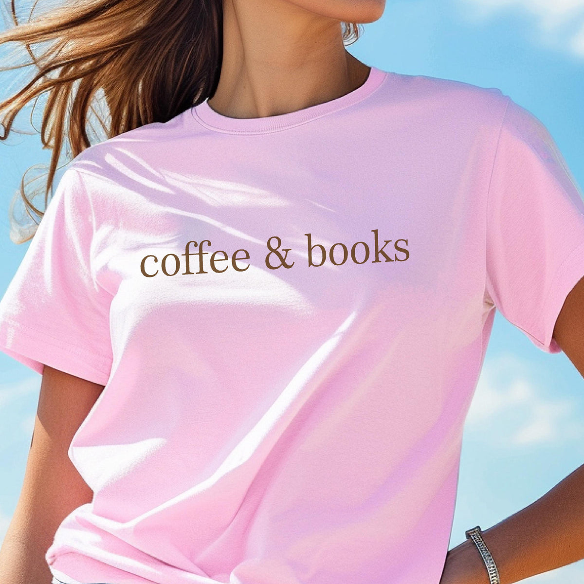 May include: A light pink short-sleeved t-shirt featuring the text "coffee & books" printed in a dark brown serif font across the chest. The garment has a classic crew neckline and a straight hem, providing a simple and clean design.
