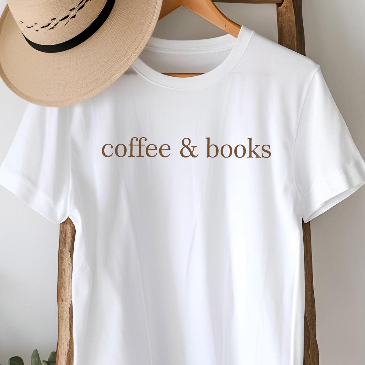 May include: A white short-sleeved t-shirt featuring the brown text coffee & books printed across the chest. The shirt is displayed alongside a tan wide-brimmed hat with a black band, both hanging on a wooden hanger.
