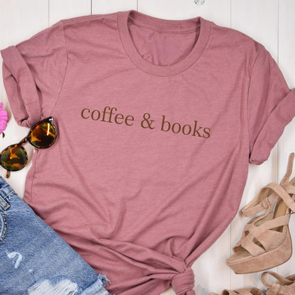 May include: A mauve short-sleeved t-shirt with the text coffee & books printed in brown across the chest. The shirt is displayed alongside a pair of tortoiseshell sunglasses, blue denim jeans, and beige heeled sandals on a white wooden surface.