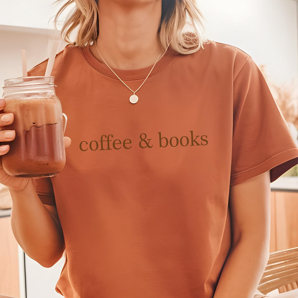 May include: A woman wearing a rust-colored t-shirt with the text "coffee & books" printed on the front. She is holding a glass of iced coffee.