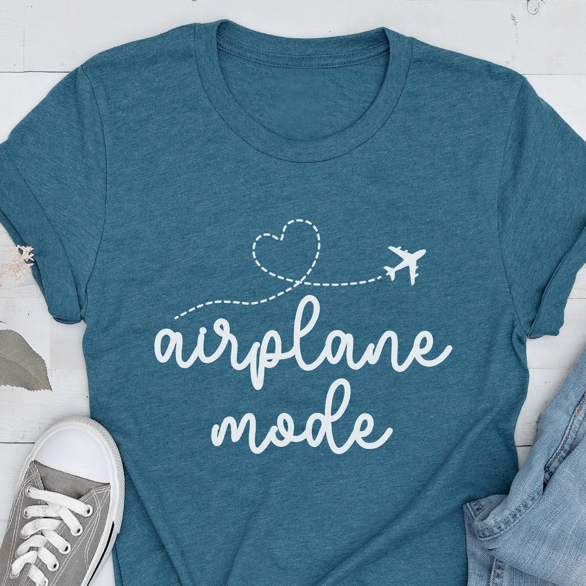 May include: Teal t-shirt with the words "airplane mode" in a white script font. A white airplane graphic follows a dotted line that forms a heart shape. A gray sneaker and denim jeans are visible in the image.