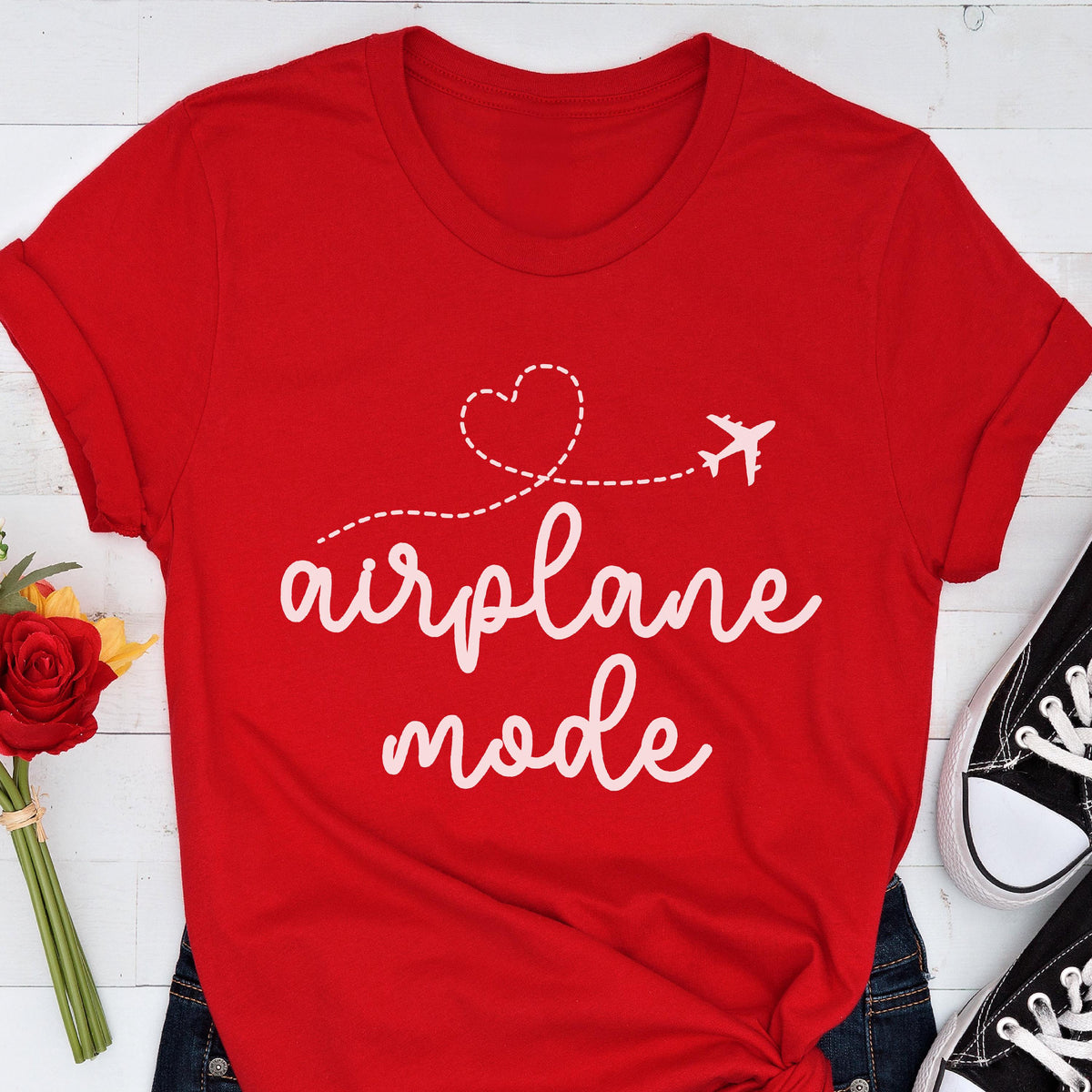 May include: A red t-shirt with the words "airplane mode" in white script. A dotted line with a heart and airplane graphic is above the text. The shirt is folded at the sleeves and is paired with blue jeans and black sneakers.