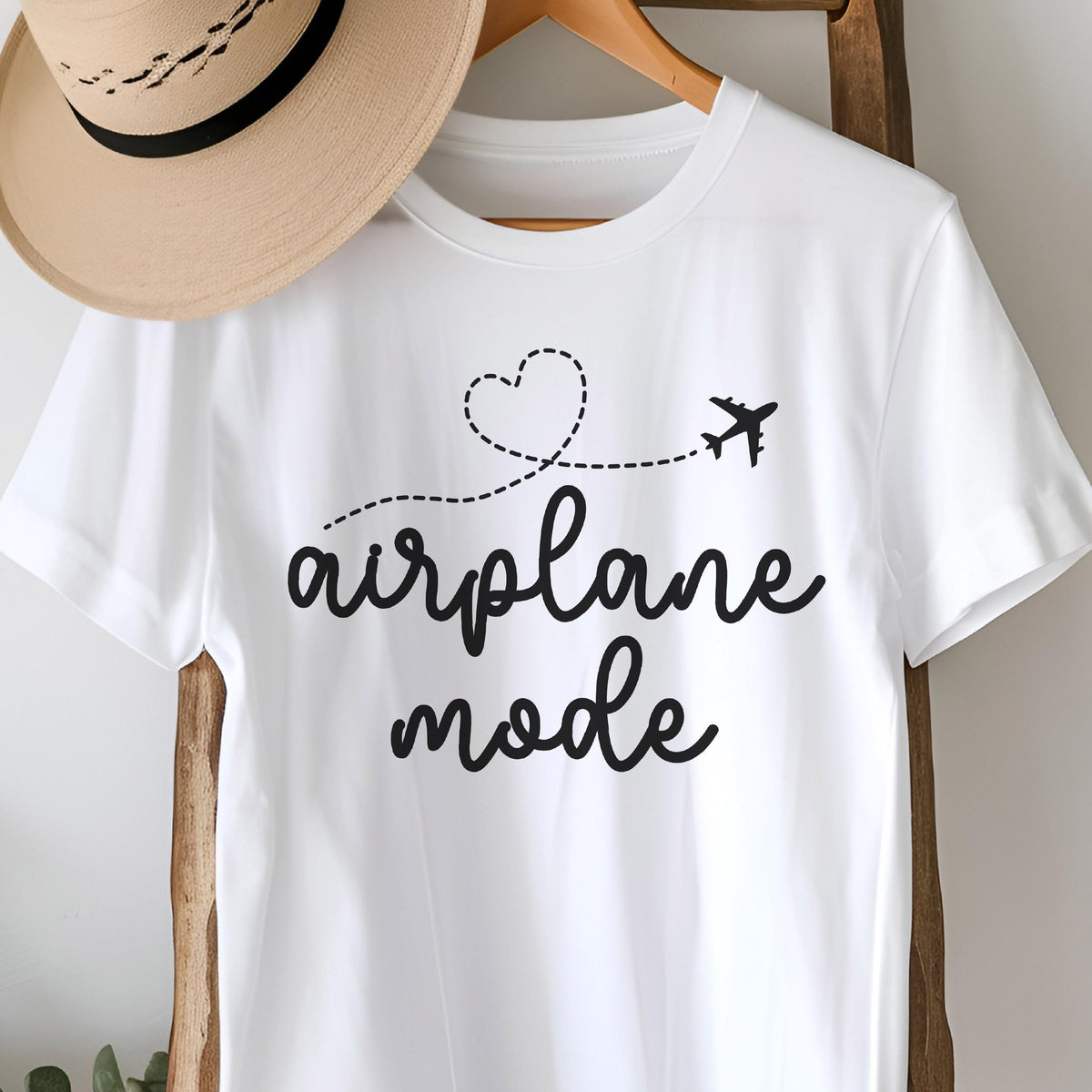 May include: White t-shirt with the words "airplane mode" in a black script font. A dotted line with a heart and airplane graphic is above the text. A tan hat hangs on a wooden hanger above the shirt.