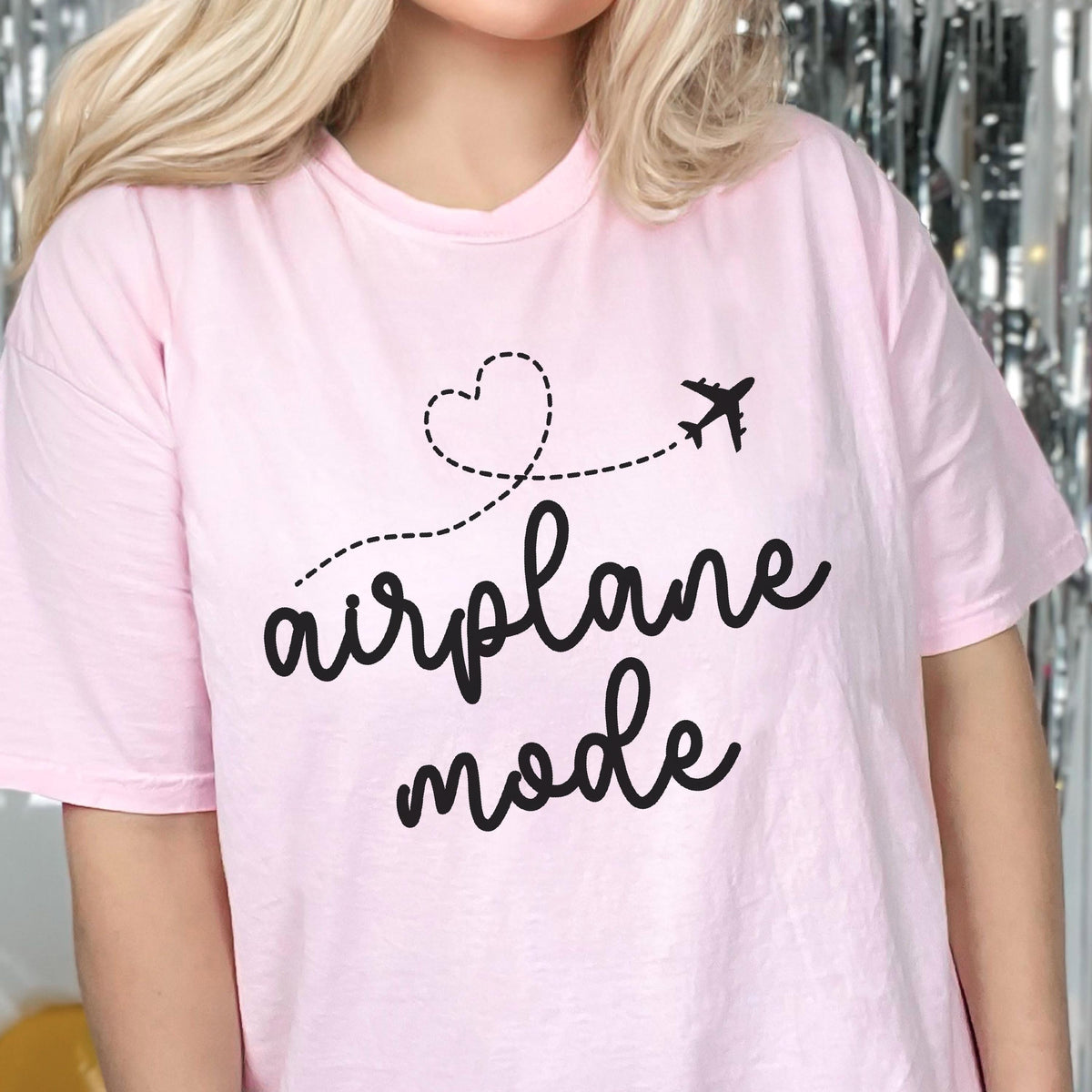 May include: A light pink t-shirt with the words "airplane mode" in a black script font. A black airplane graphic follows a dotted line that forms a heart shape. The shirt is a casual style.