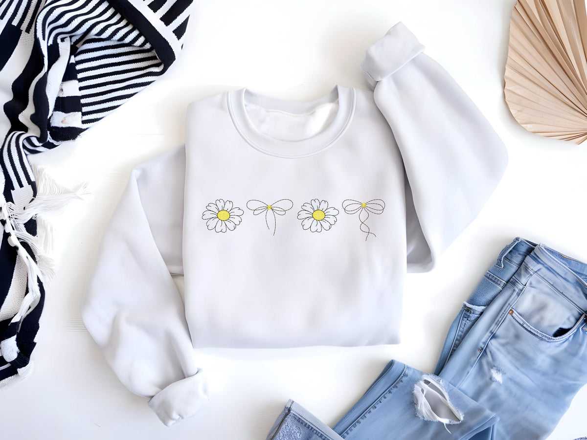 May include: A white sweatshirt with a simple design of four daisies with yellow centers and a bow tied around each stem. The sweatshirt is laid flat on a white surface.
