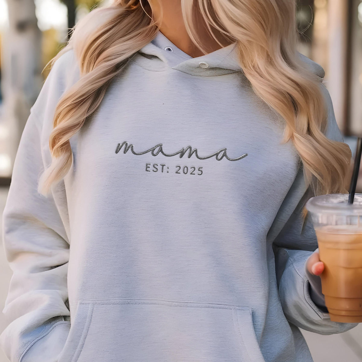 May include: A light blue hooded sweatshirt featuring the embroidered text mama and EST: 2025 in a dark gray script and sans-serif font. The garment includes a front kangaroo pocket, a drawstring hood with metal eyelets, and long sleeves.