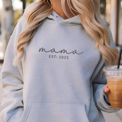 May include: A light blue hooded sweatshirt featuring the embroidered text mama and EST: 2025 in a dark gray script and sans-serif font. The garment includes a front kangaroo pocket, a drawstring hood with metal eyelets, and long sleeves.
