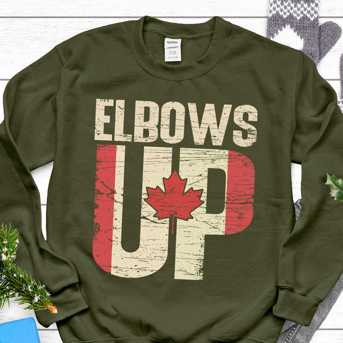 May include: A green sweatshirt with the text "Elbows Up" in white letters. The text is overlaid with a distressed Canadian flag design.