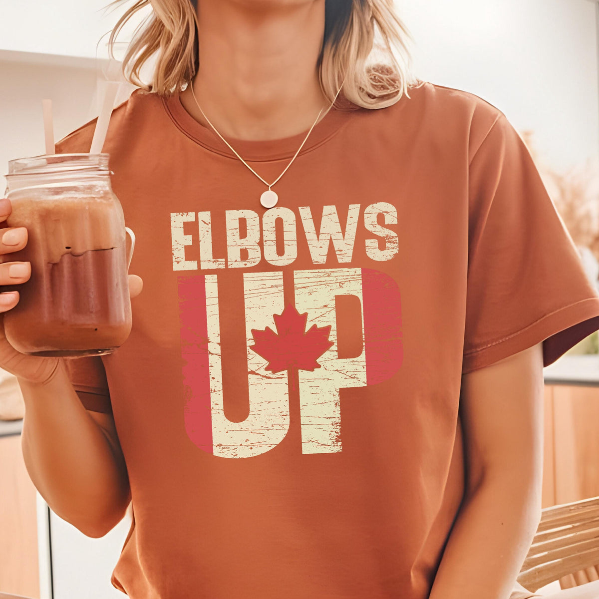 May include: A brown t-shirt with the text "Elbows Up" in white with a red maple leaf inside the "U".