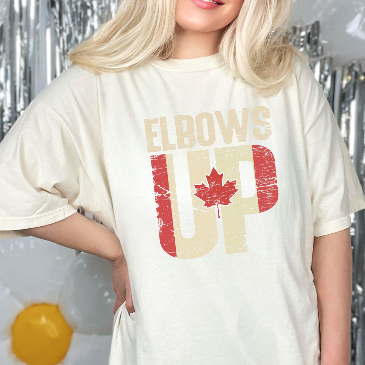 May include: A white t-shirt with a distressed graphic design featuring the text "Elbows Up" and a red maple leaf in the shape of a Canadian flag.
