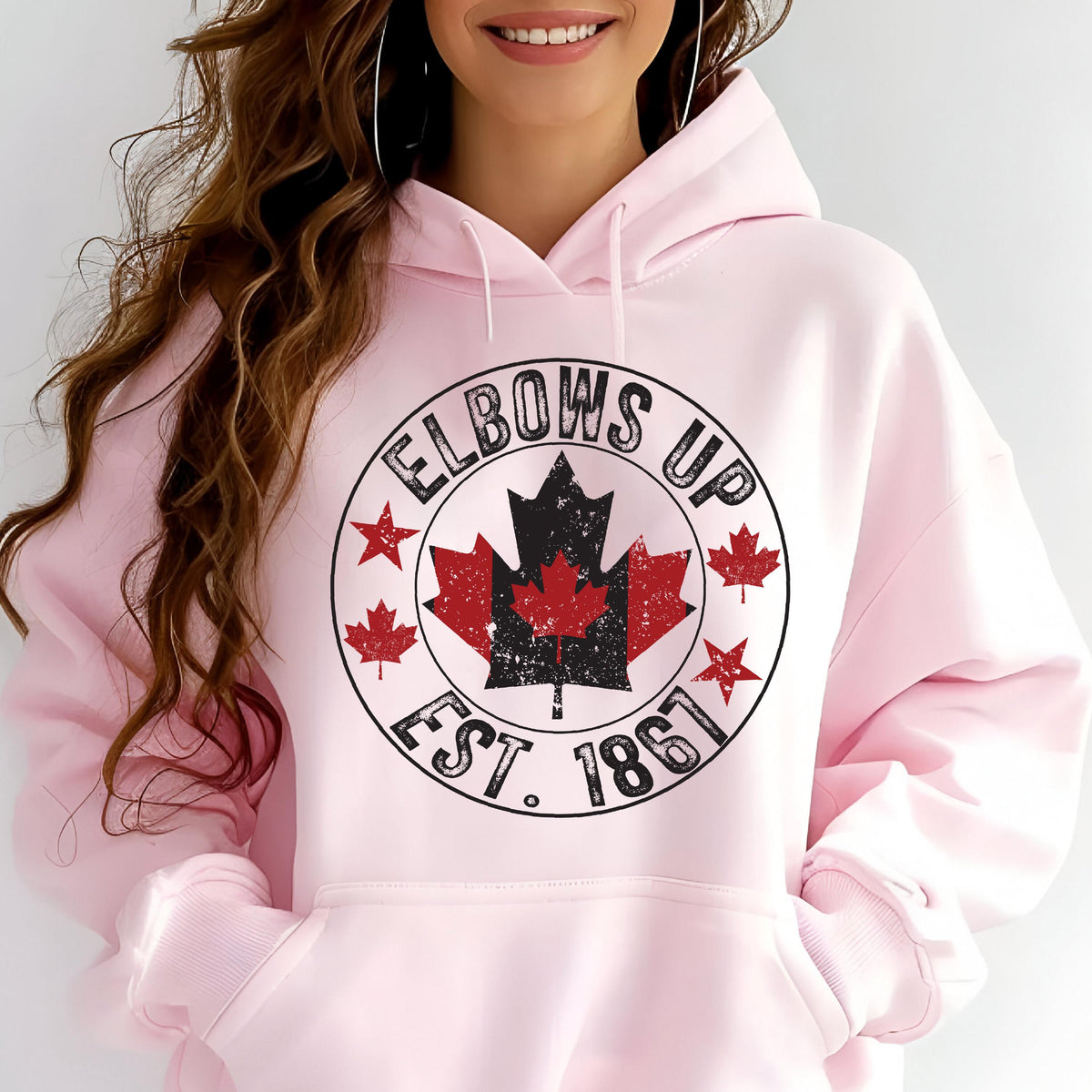 May include: A light pink hooded sweatshirt featuring a central circular graphic with the text ELBOWS UP EST. 1867. The design includes a large distressed black and red maple leaf, smaller red maple leaves, and red stars within the black circular border.