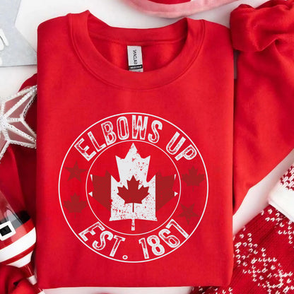May include: A bright red crewneck sweatshirt featuring a white circular graphic with the text ELBOWS UP EST. 1867. The design includes a central white maple leaf with a distressed texture, surrounded by smaller red maple leaf and star silhouettes.