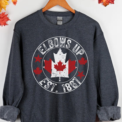 May include: A dark heather gray long-sleeve sweatshirt featuring a circular graphic with the text ELBOWS UP and EST. 1867. The center contains a large white and red maple leaf design, surrounded by smaller red maple leaf icons within the circle.