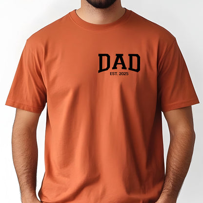 May include: A burnt orange t-shirt with the text "DAD EST. 2025" printed on the front.