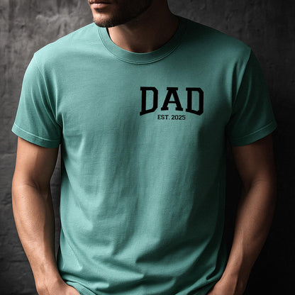 May include: A teal blue t-shirt with the word "DAD" in black block letters and the text "EST. 2025" underneath.