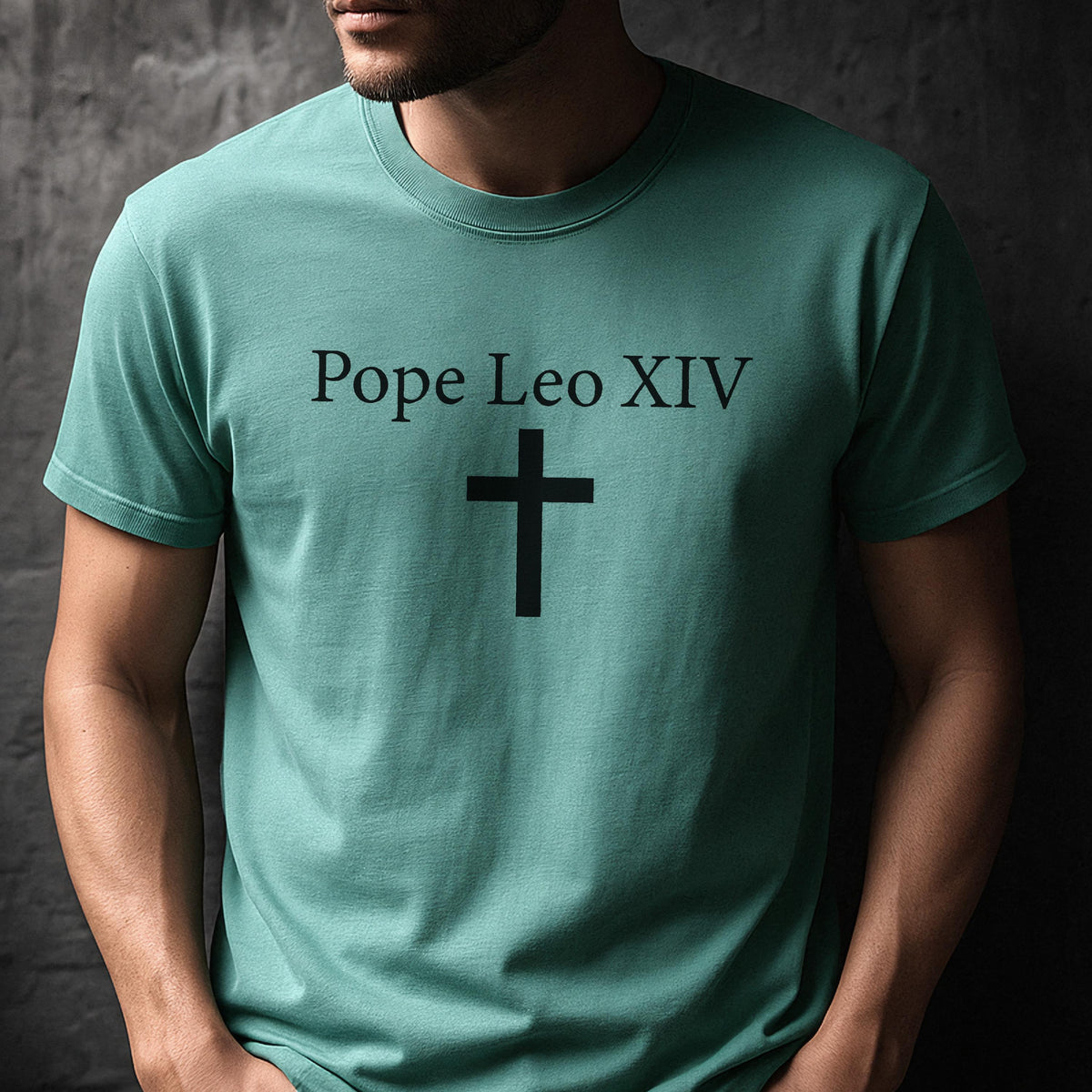 May include: A teal-colored t-shirt with the text "Pope Leo XIV" above a black cross. The shirt is made of a soft material and has short sleeves. The design is simple and centered on the chest.