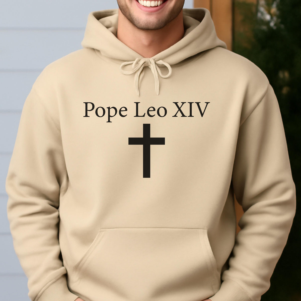 May include: A beige hooded sweatshirt with the text "Pope Leo XIV" above a black cross. The sweatshirt has a drawstring at the neckline and a front pocket.