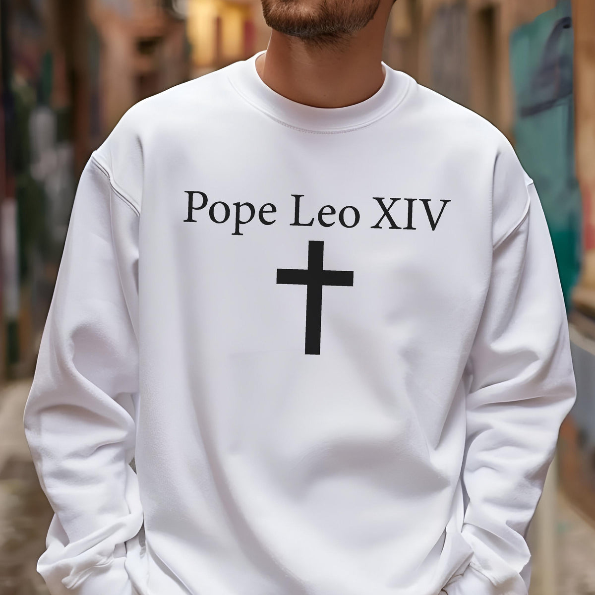 May include: White sweatshirt with the text "Pope Leo XIV" above a black cross. The sweatshirt has a crew neck and long sleeves. The image is taken from the chest up.