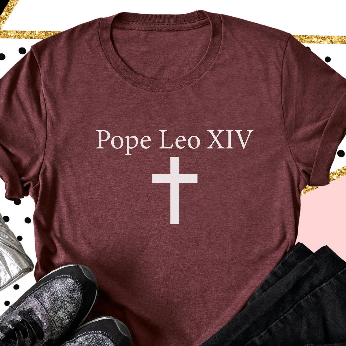 May include: A maroon t-shirt with the text "Pope Leo XIV" above a white cross. The shirt has a crew neck and short sleeves. The image is set against a decorative background with gold and black accents.