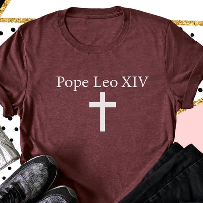 May include: A maroon t-shirt with the text "Pope Leo XIV" above a white cross. The shirt has a crew neck and short sleeves. The image is set against a decorative background with gold and black accents.