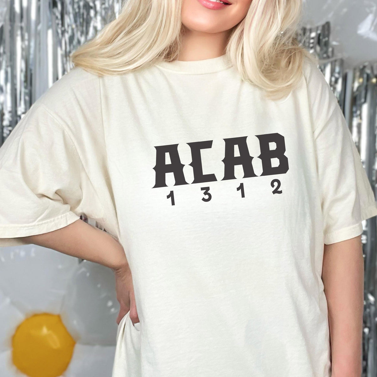 May include: An off-white short-sleeve t-shirt featuring the black text ACAB printed in a bold, stylized font across the chest. Below the letters, the numbers 1 3 1 2 are printed in a matching black font, centered horizontally on the front of the shirt.