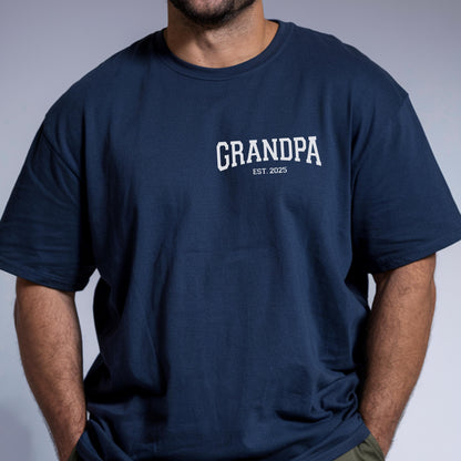 May include: Navy blue t-shirt with the word "GRANDPA" in white, arched text. Below the word "GRANDPA" is the text "EST. 2025". The shirt is a solid color and has short sleeves.