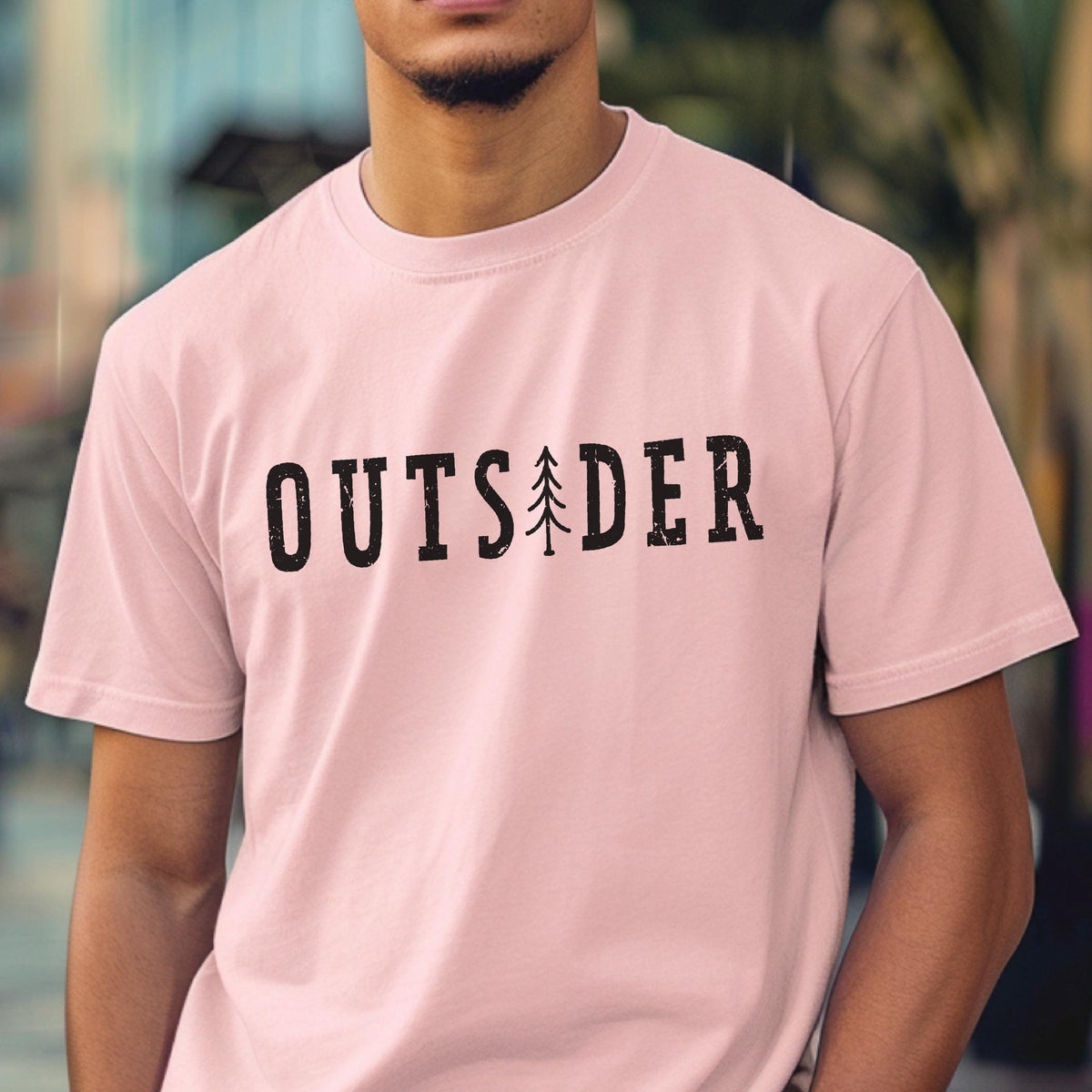 May include: A light pink short-sleeved t-shirt featuring the word OUTSIDER printed in black, distressed capital letters across the chest, with a stylized black pine tree graphic replacing the letter I in the center of the word.