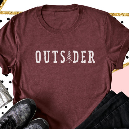 May include: A maroon short-sleeved t-shirt featuring the white text "OUTSIDER" printed across the chest, with a stylized white pine tree graphic replacing the letter "I" in the word. The fabric has a heathered texture and a crew neckline.