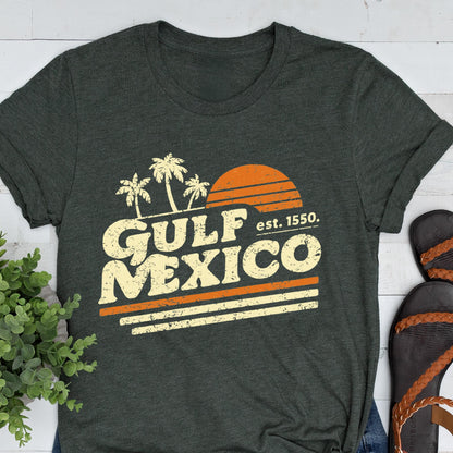 May include: Dark gray t-shirt featuring a retro design with the words "GULF MEXICO" in a vintage font, palm trees, and a setting sun. The text "est. 1550" is also included. The shirt has rolled-up sleeves and is paired with sandals.