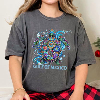 May include: A charcoal gray short-sleeve t-shirt featuring a vibrant, multi-colored sea turtle graphic with intricate floral patterns on its shell. The design includes swirling blue water accents and the text GULF OF MEXICO in light blue letters.