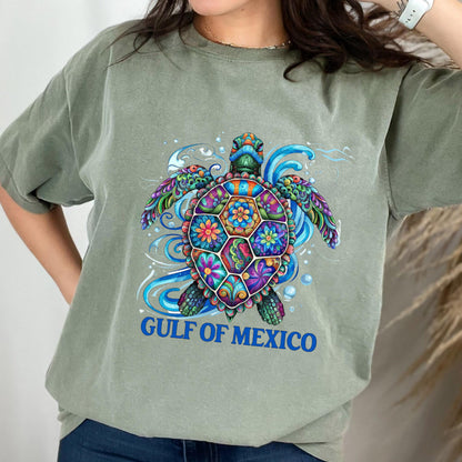 May include: A sage green short-sleeve t-shirt featuring a colorful, patterned sea turtle graphic with blue swirling water elements. Below the turtle, the text GULF OF MEXICO is printed in bold blue capital letters on the front of the garment.