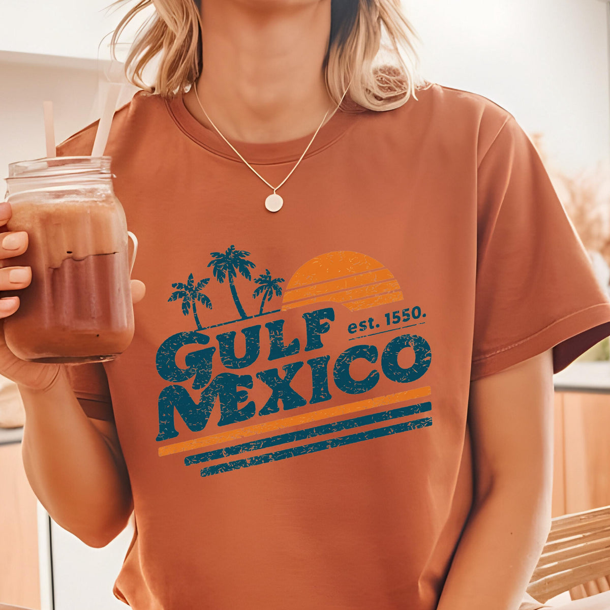 May include: A rust-colored t-shirt featuring a graphic design with the text "GULF MEXICO" in teal, palm trees, and a setting sun. The text "est. 1550" is also included in the design. The shirt is short-sleeved.