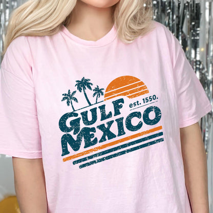 May include: A light pink t-shirt featuring a graphic design with the words "GULF MEXICO" in teal, palm trees, and an orange sun. The text "est. 1550." is also included. The shirt is a casual wear item.