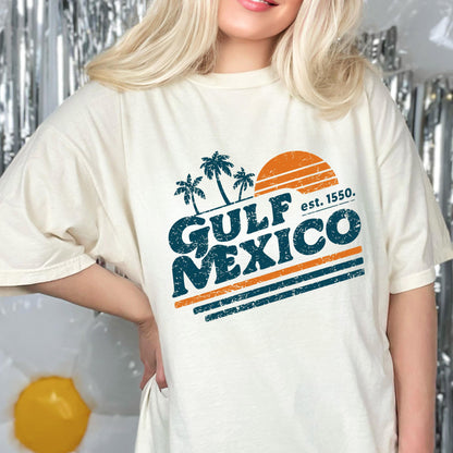 May include: Cream-colored t-shirt featuring a distressed graphic design. The design includes palm trees, a setting sun, and the words "GULF MEXICO" in teal, with the text "est. 1550." in white and orange stripes.