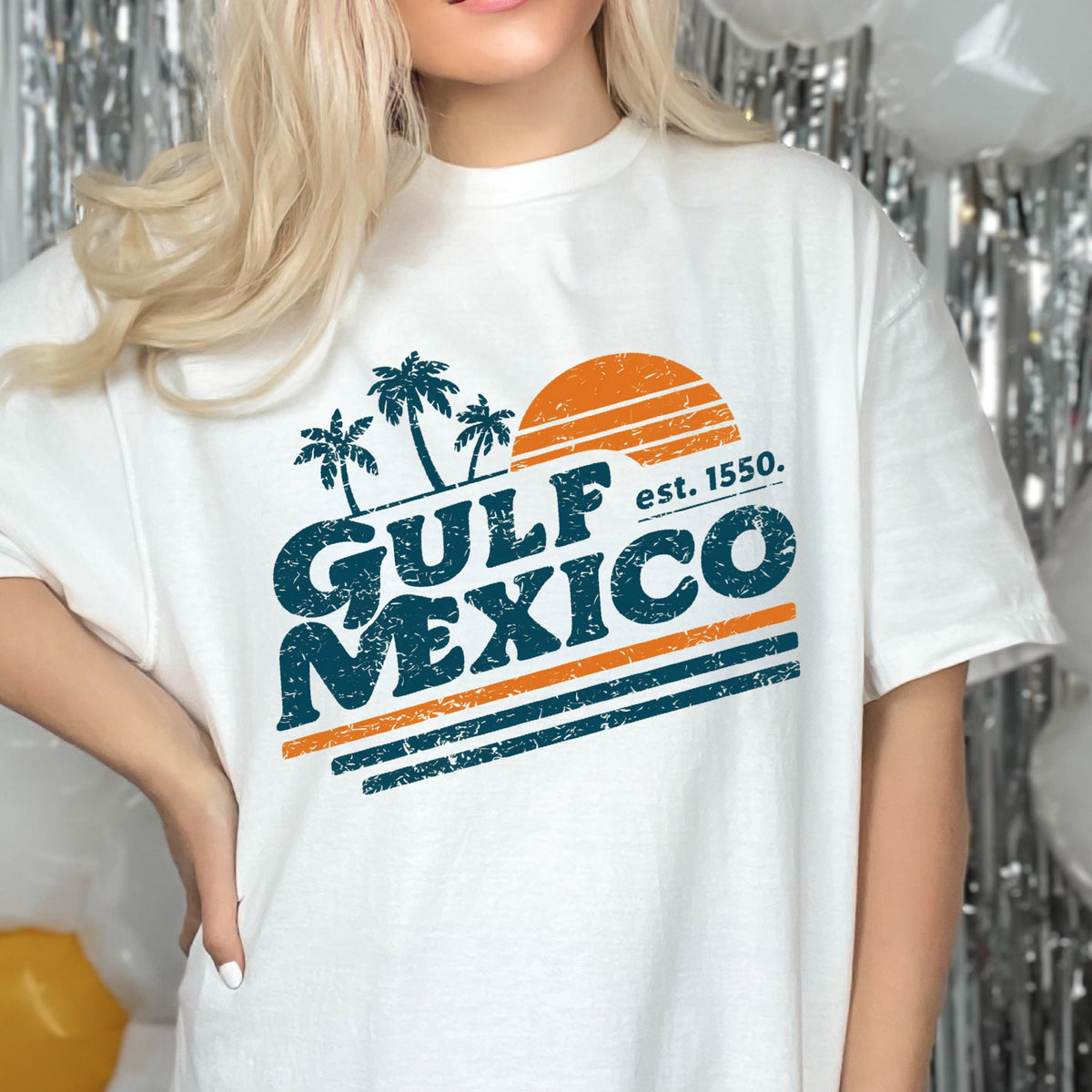 May include: White t-shirt with a retro design featuring the text "GULF MEXICO" in teal and orange, with palm trees and a sun graphic. The text "est. 1550." is also included. A casual, beach-themed apparel item.