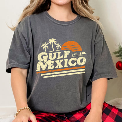 May include: Dark gray t-shirt featuring a retro design with the words "GULF MEXICO" in a vintage font, palm trees, and a setting sun graphic. The text "est. 1550" is also included. The shirt has a relaxed fit.