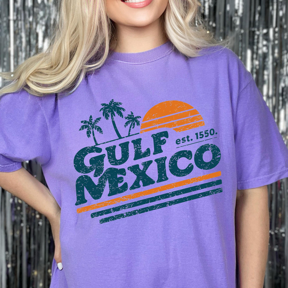 May include: A purple t-shirt featuring a graphic design with the words "GULF MEXICO" in teal, palm trees, and an orange sun. The text "est. 1550" is also included in the design. The shirt is a comfortable, casual style.
