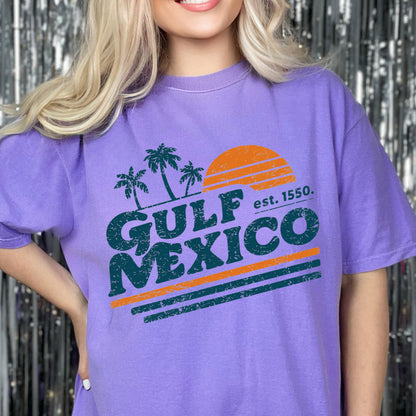 May include: A purple t-shirt featuring a graphic design with the words "GULF MEXICO" in teal, palm trees, and an orange sun. The text "est. 1550" is also included in the design. The shirt is a comfortable, casual style.