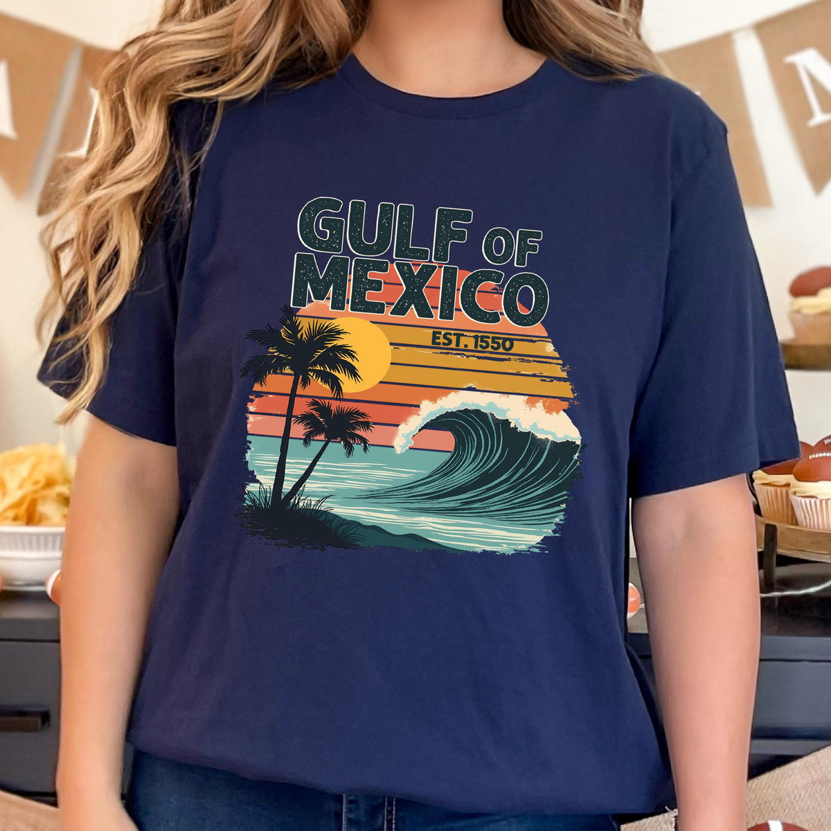 May include: A navy blue t-shirt with a retro-style graphic of a wave crashing on a beach with palm trees and a sunset. The text "GULF OF MEXICO EST. 1550" is printed on the shirt.