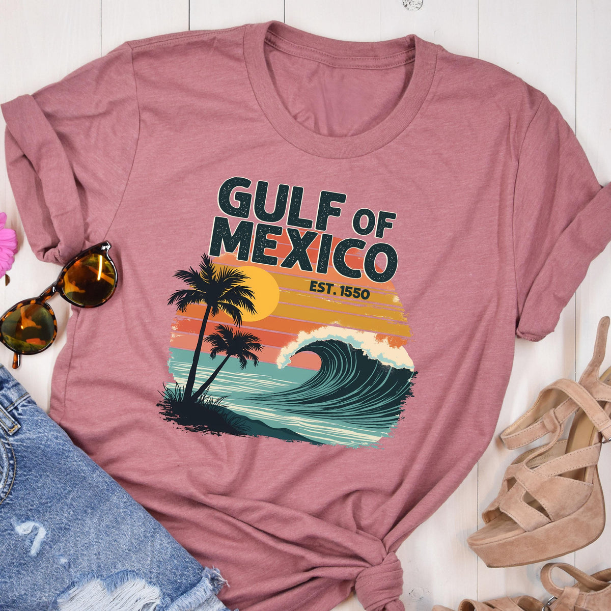 May include: A pink t-shirt with a retro-style graphic design featuring a wave, palm trees, and the text "GULF OF MEXICO EST. 1550".