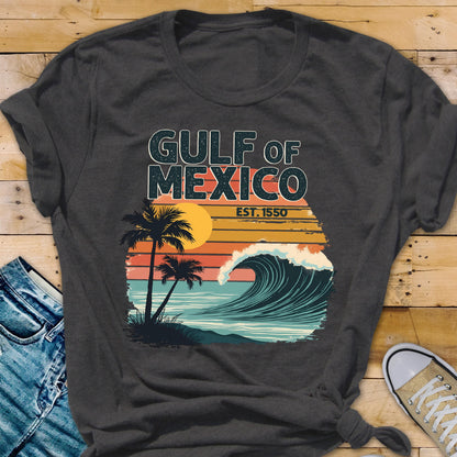 May include: A dark gray t-shirt with a vintage-style graphic of a wave crashing on a beach with palm trees and the text "GULF OF MEXICO EST. 1550".