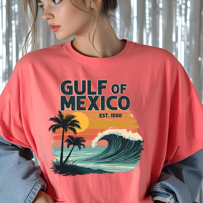 May include: A pink t-shirt with a graphic design of a sunset over a wave with palm trees. The text "GULF OF MEXICO EST. 1550" is printed on the shirt.