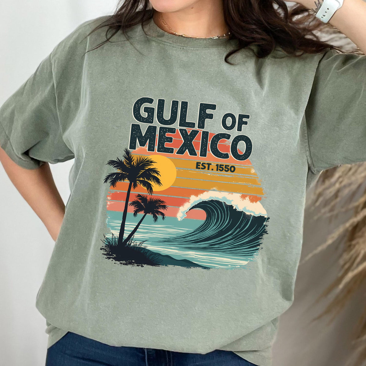 May include: A green t-shirt with a vintage-style graphic of a wave, palm trees, and a sunset. The text "GULF OF MEXICO EST. 1550" is printed on the shirt.